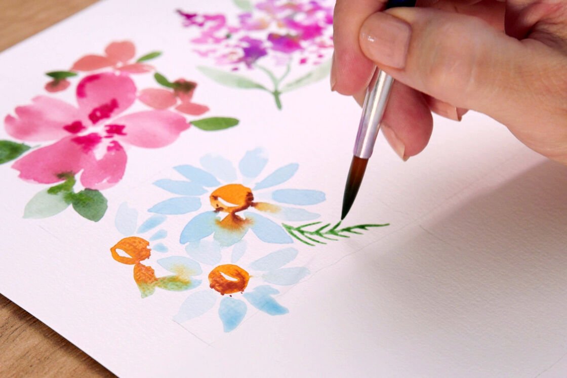uma mão desenha várias flores com tintas de aguarela
